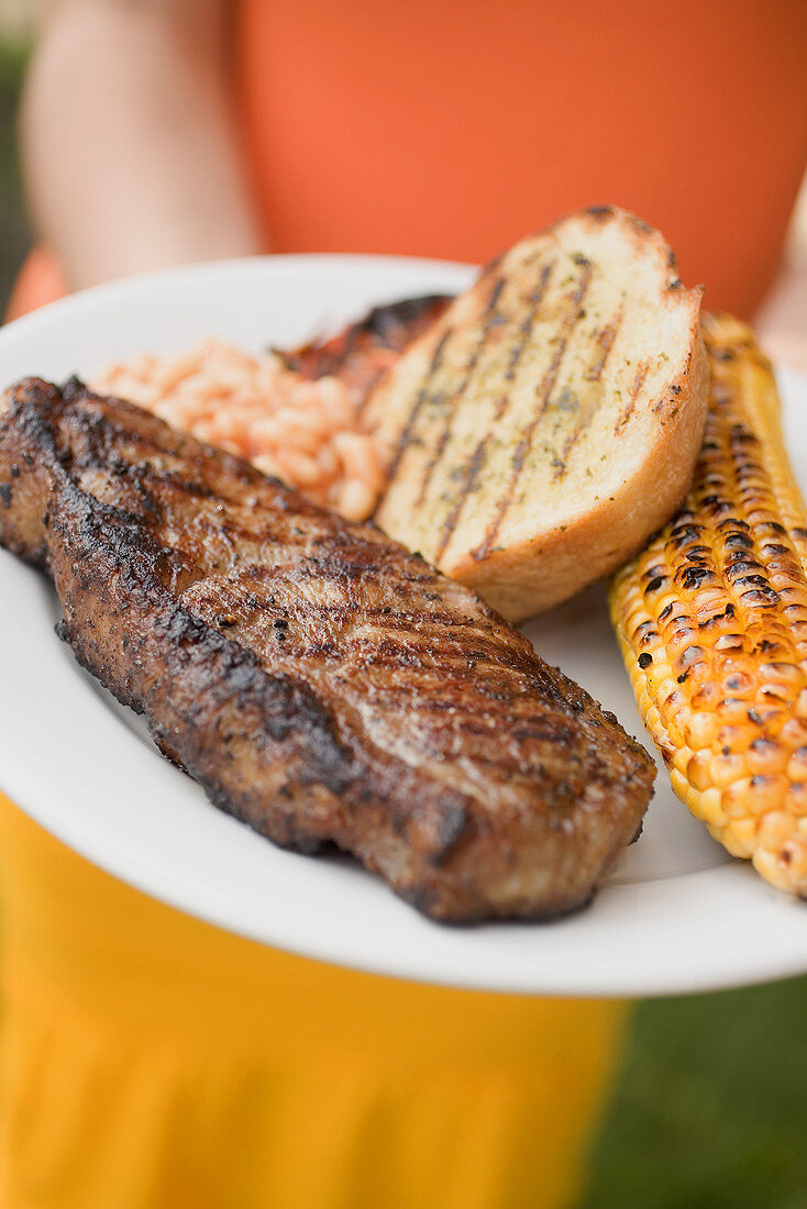 Woman holding a plate of beef steak, corn on the cob, toast
