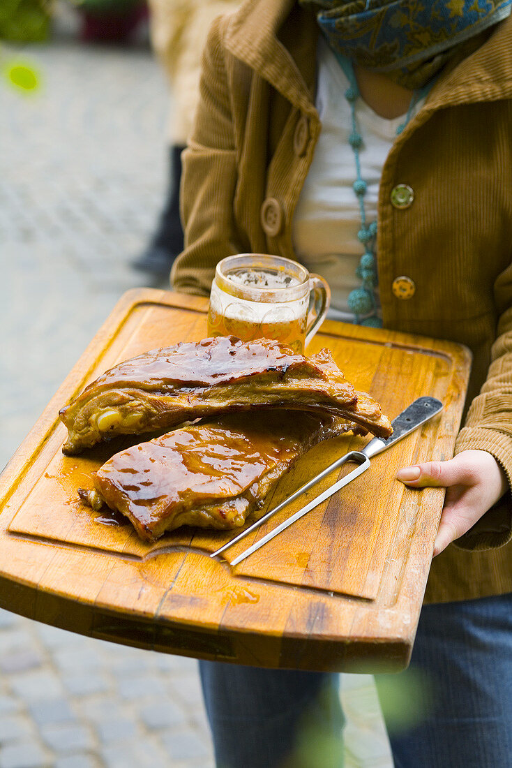 Frau hält Schneidebrett mit glasierten Rippchen und Glas Bier