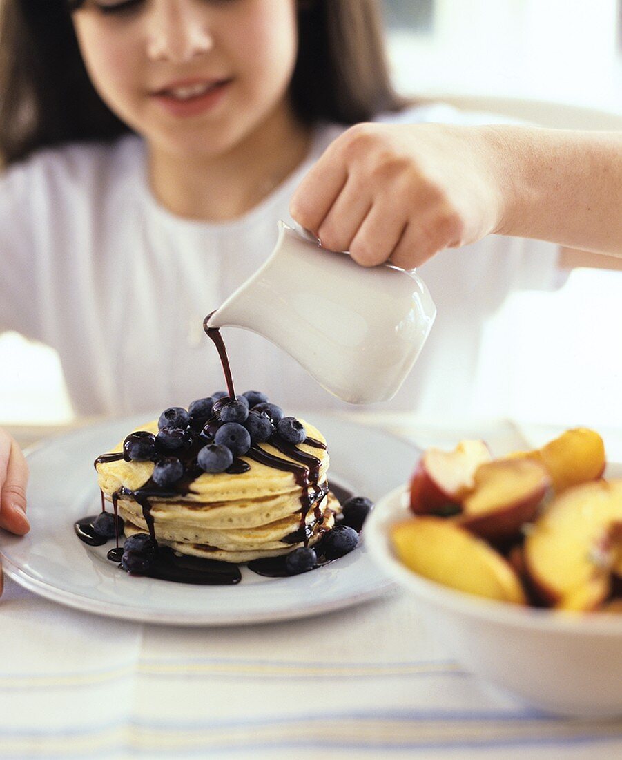 Girl pouring chocolate sauce over pancakes and blueberries