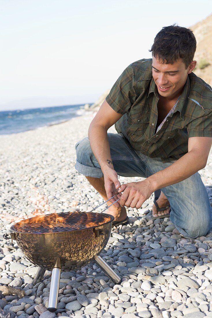 Junger Mann kniet vor brennendem Grill am Strand
