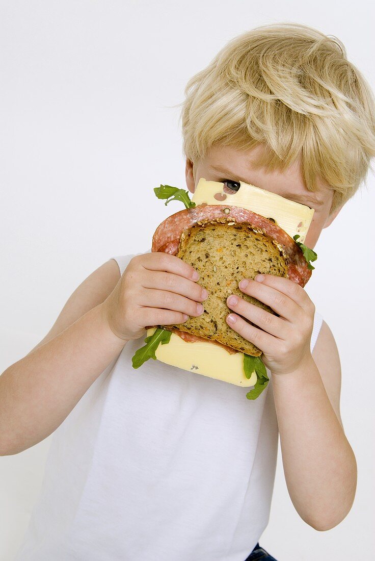 Blonder Junge hält Salami-Käse-Brot vors Gesicht