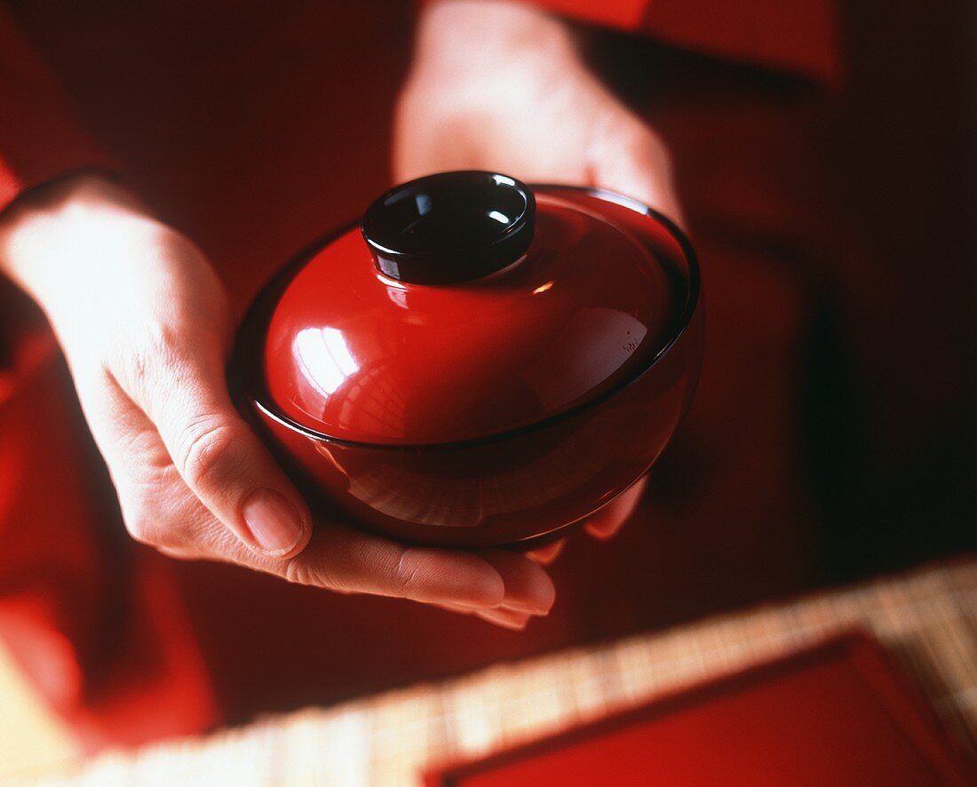 Hands holding a red soup bowl with a lid