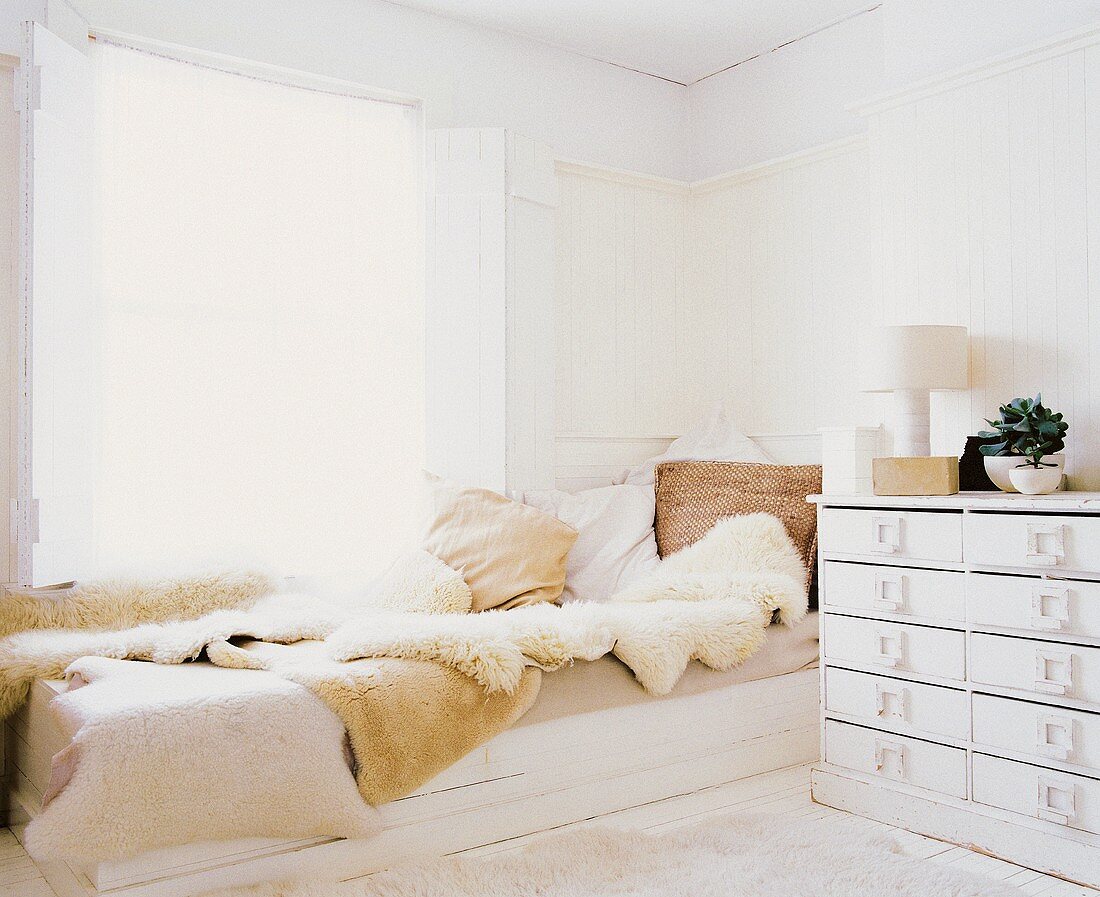 Bett mit Lammfellen und mehreren Kissen aus Naturmaterialien; daneben eine weiße Schubladenkommode im Shabby Stil