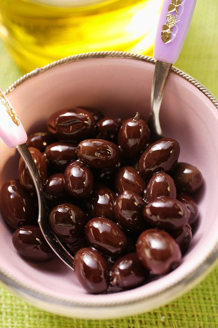 Eingelegte schwarze Oliven im Schälchen, dahinter Olivenöl