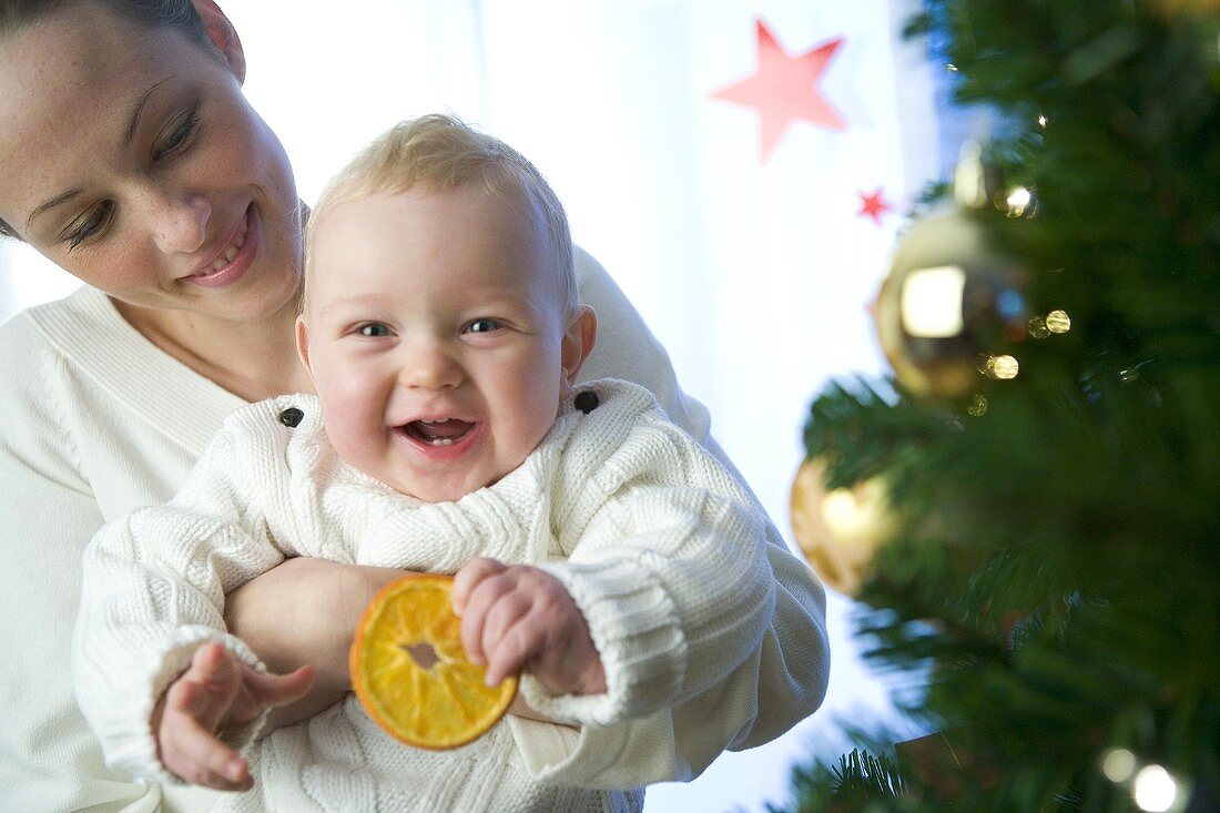 Mutter hält Baby mit Orangenscheibe vor Weihnachtsbaum