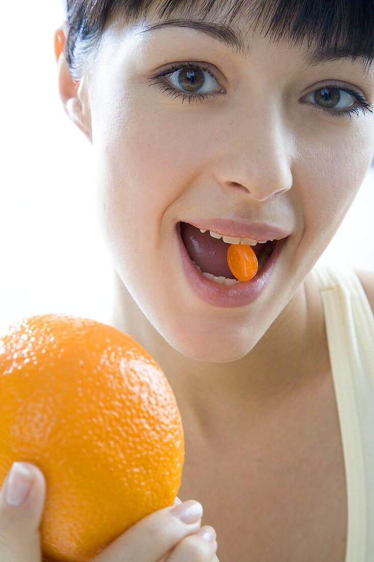 Frau hält orangefarbenes Dragee im Mund & Orange in der Hand
