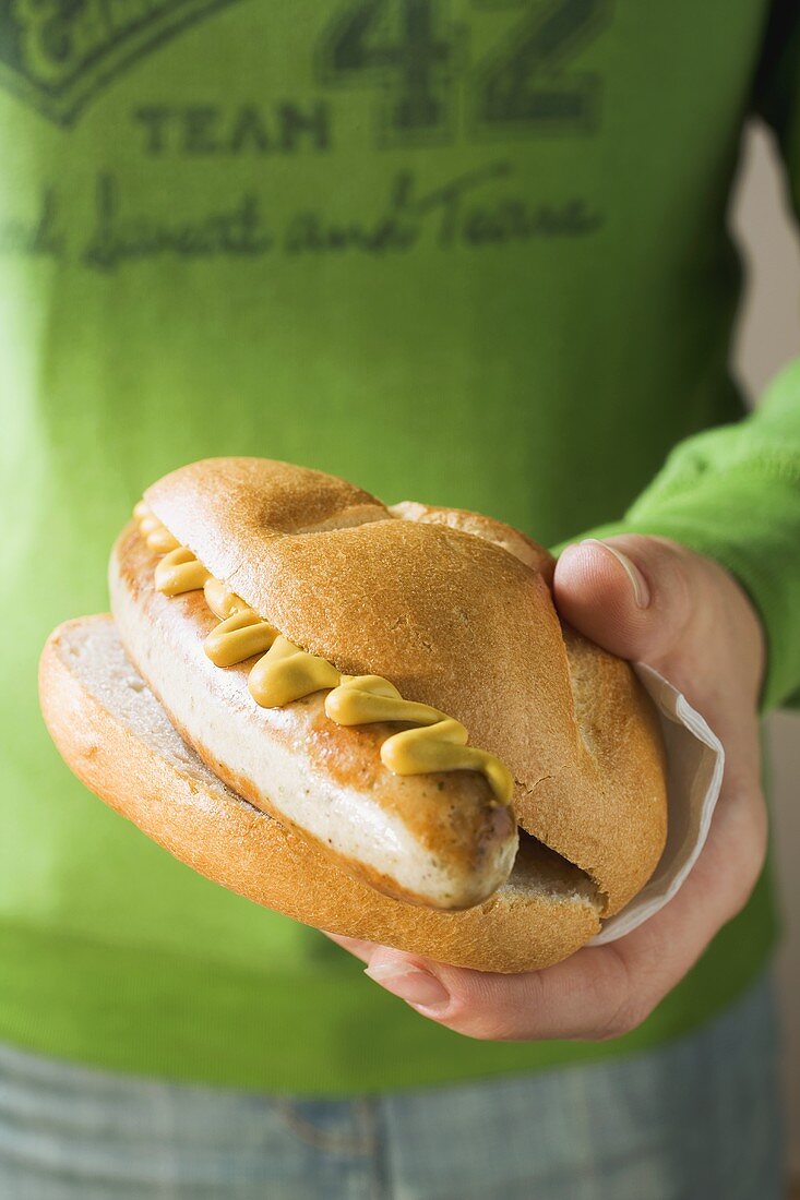 Hand holding sausage and mustard in a bread roll