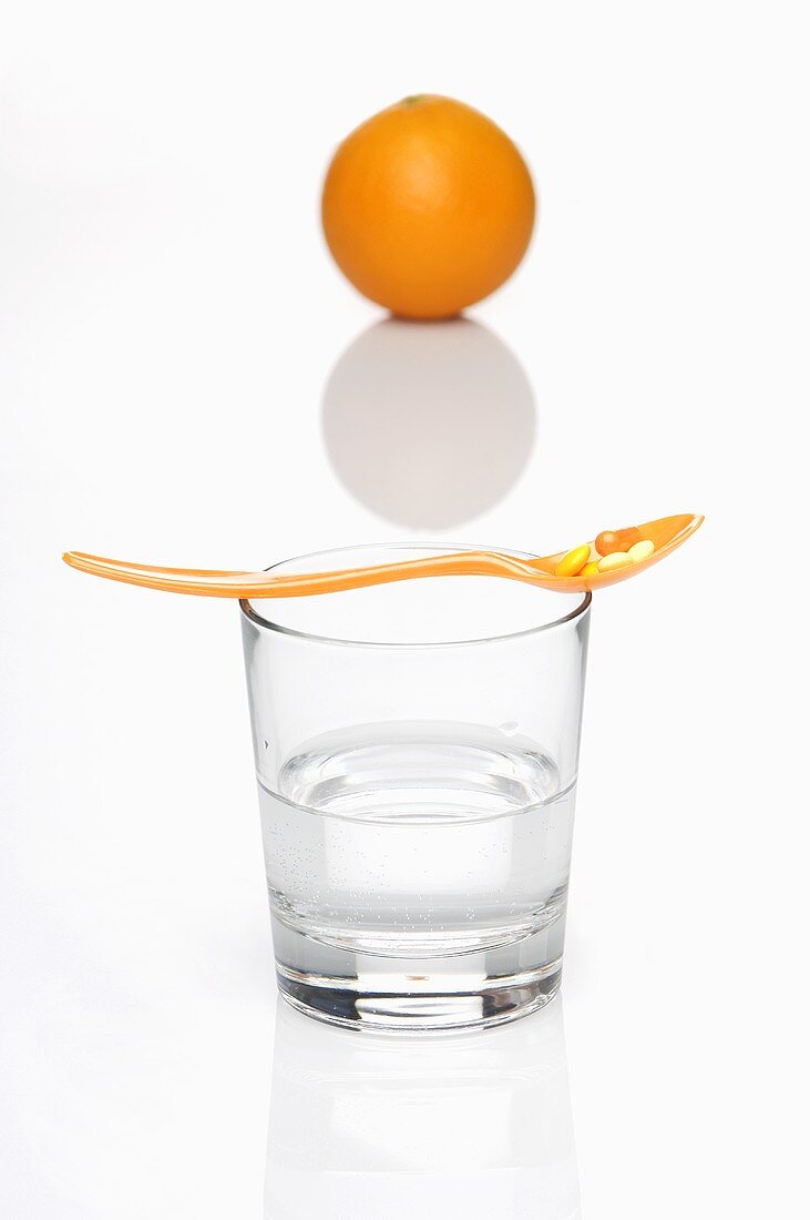 Vitamin tablets on a spoon on top of a glass of water with an orange in the background