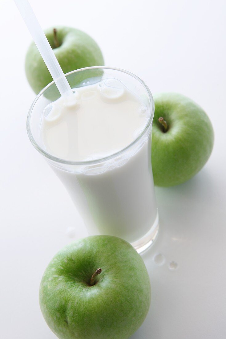 A glass of milk with a straw and three green apples