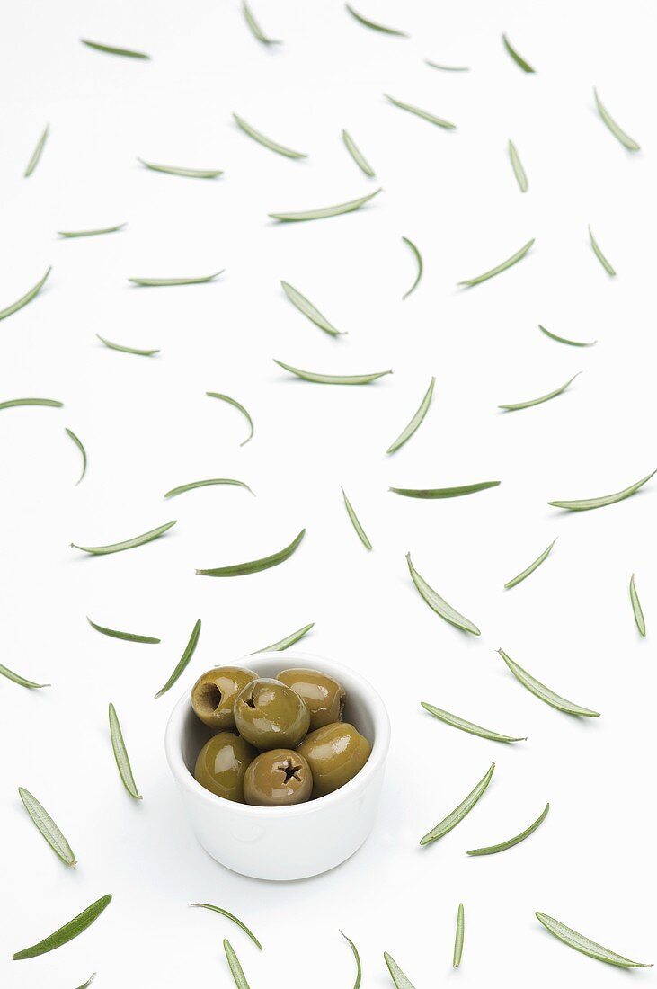 Green olives in a bowl and scattered rosemary leaves