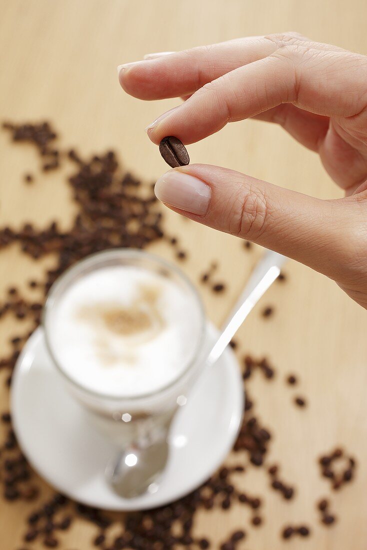 Hand hält eine Kaffeebohne über einem Glas Latte Macchiato
