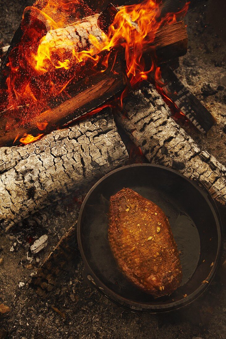 Gewürzter Lammbraten in Pfanne neben Holzfeuer