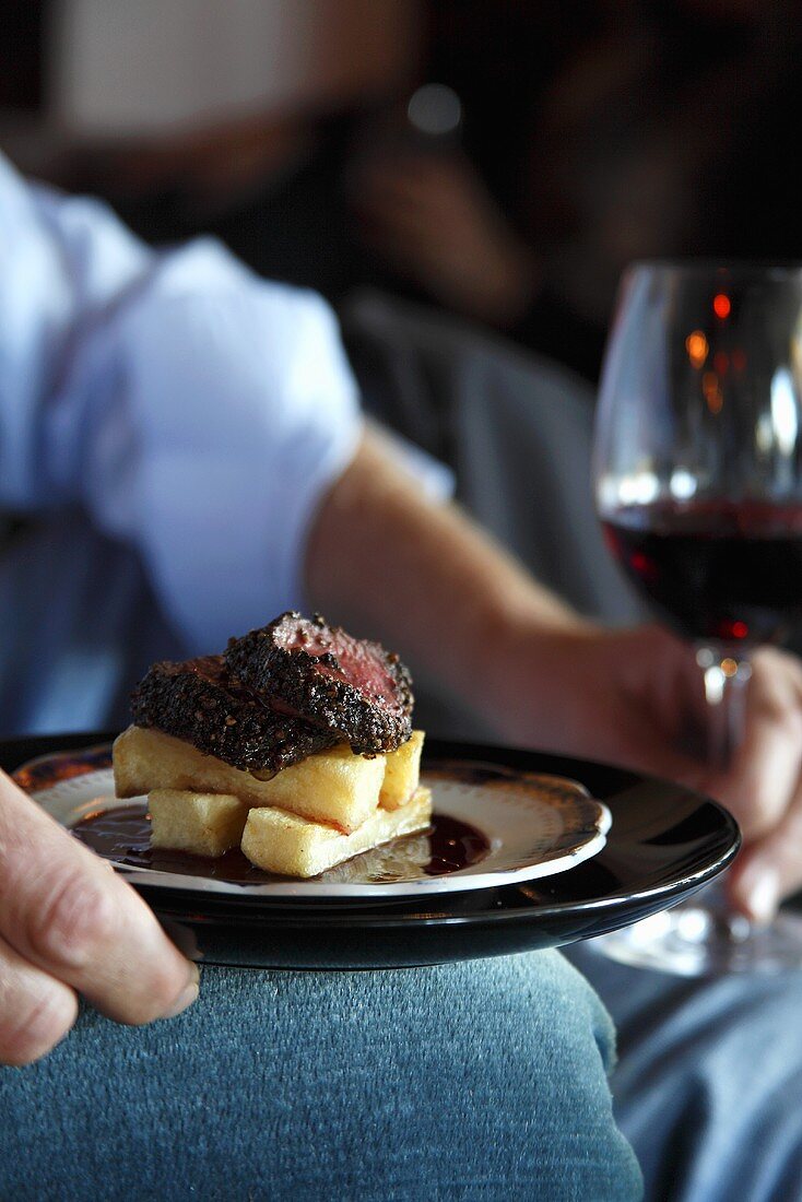 A man holding a plate of ostrich steak and chips and a glass of red wine