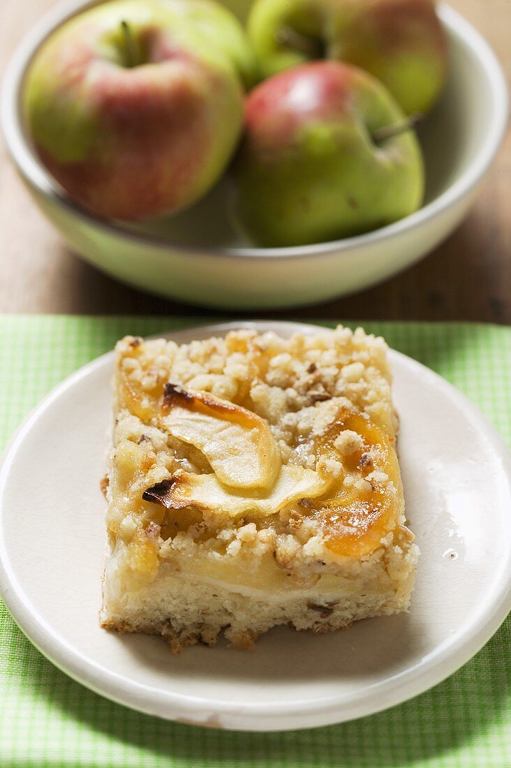 Stück Apfel-Streusel-Kuchen, frische Äpfel im Hintergrund