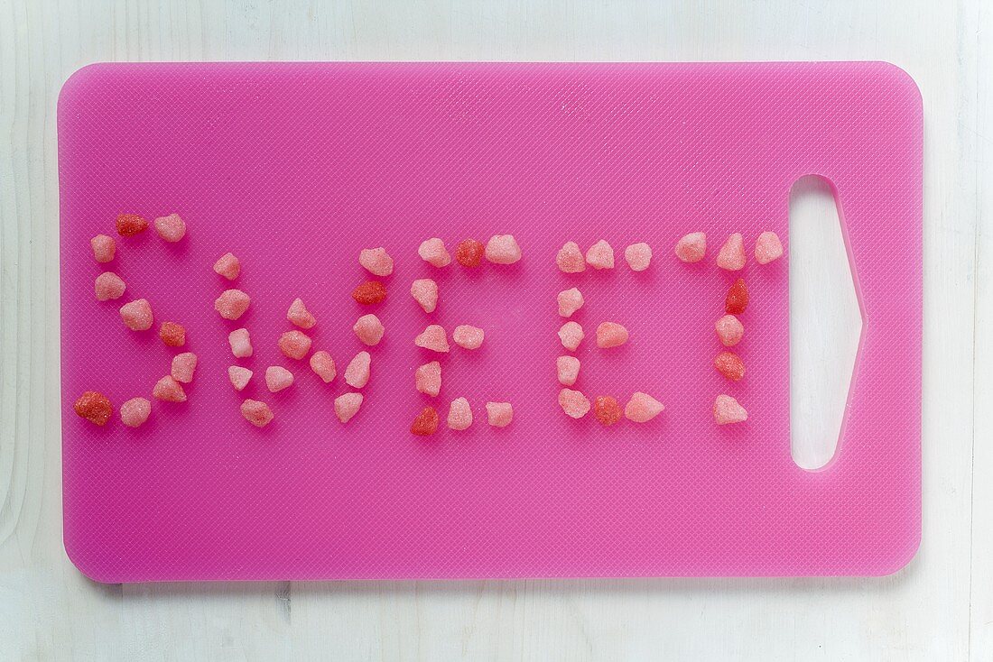 Schriftzug 'Sweet' aus Zuckerbonbons auf rosa Schneidebrett