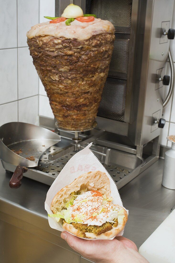 Hand hält aufgeklappten Döner Kebab vor Drehspiess