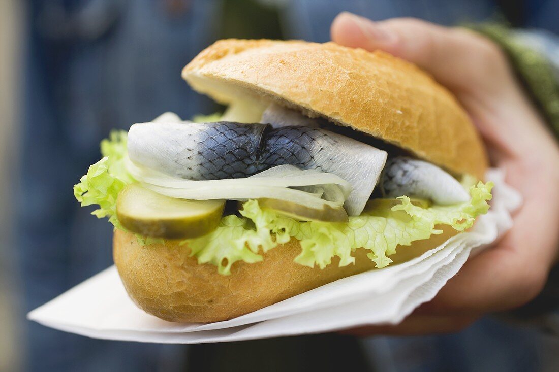 Hand holding bread roll filled with herring, onions & gherkins