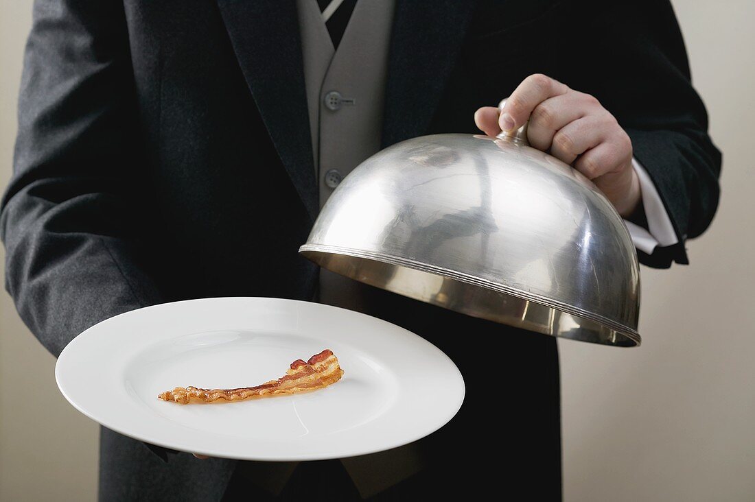 Butler serving rasher of fried bacon on plate with dome cover