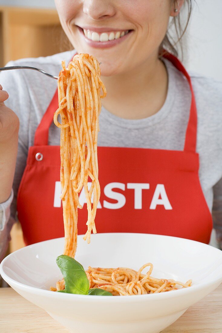 Young woman in apron eating spaghetti with tomato sauce