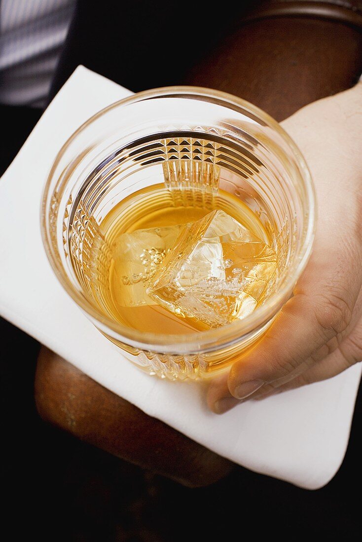 Hand holding glass of whisky with ice cubes