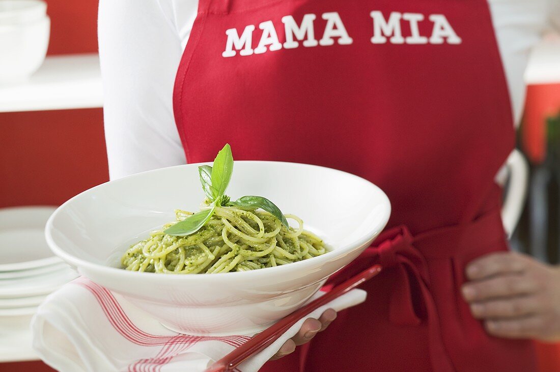 Frau mit roter Schürze hält Teller Spaghetti mit Pesto