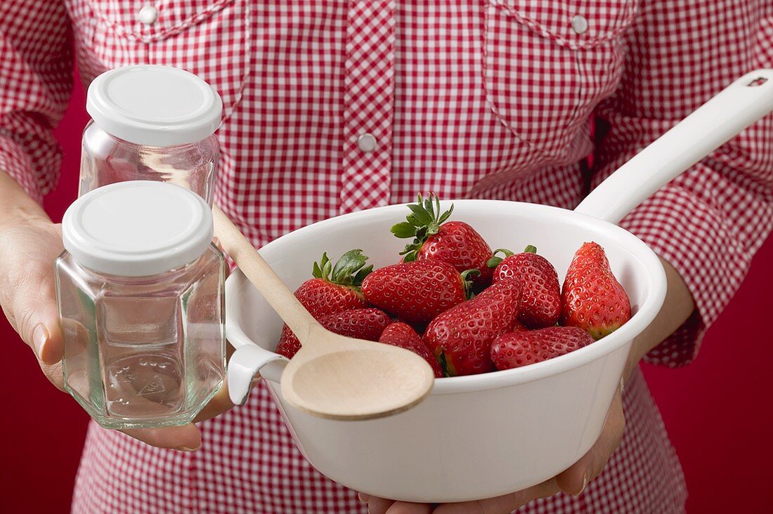 Frau hält Seiher mit Erdbeeren, Marmeladengläser und Kochlöffel