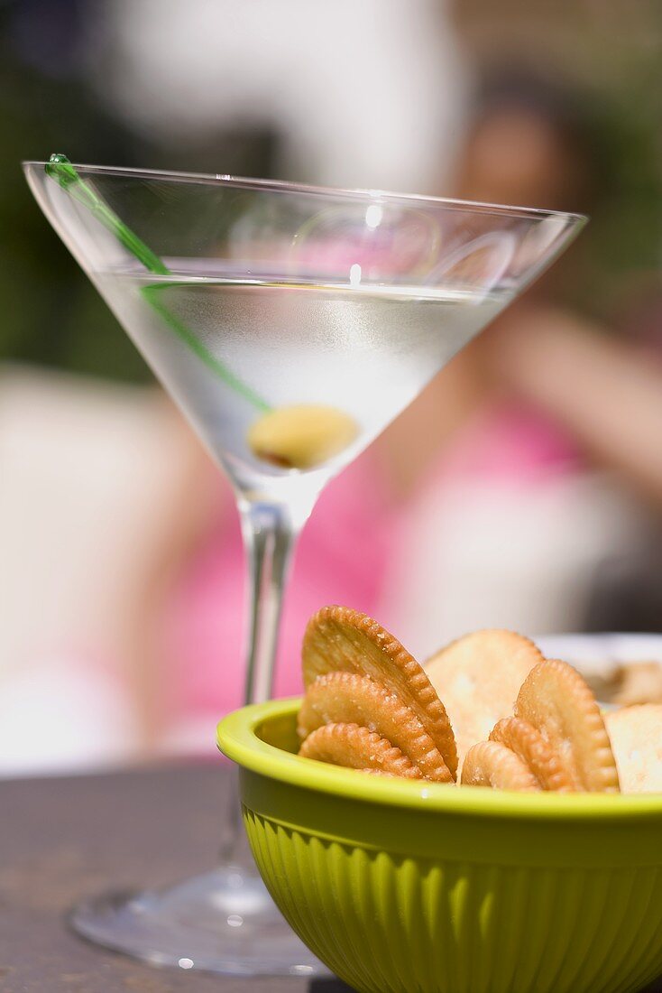 Martini with green olive, crackers, woman in background