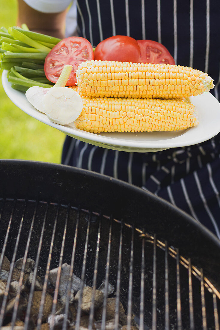 Person holding plate of vegetables & corn on the cob for grilling