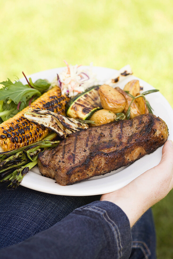 Frau hält Teller mit Steak, Grillgemüse und Maiskolben