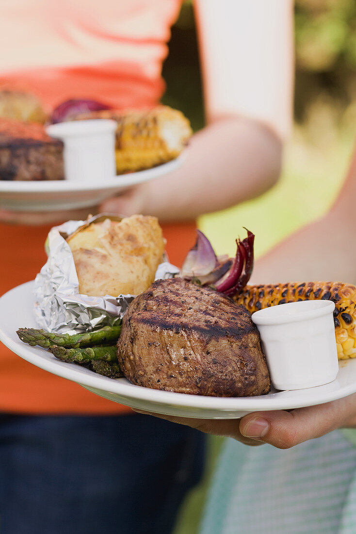 Two women holding plates of grilled steak & accompaniments