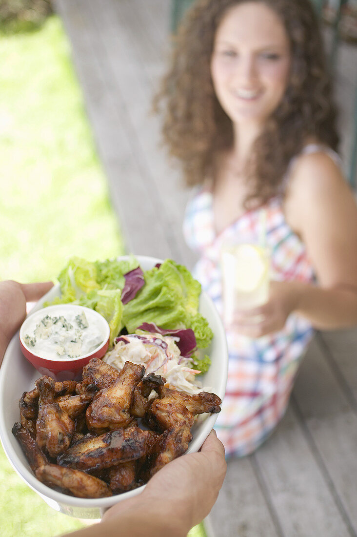 Hands serving grilled chicken wings, woman in background