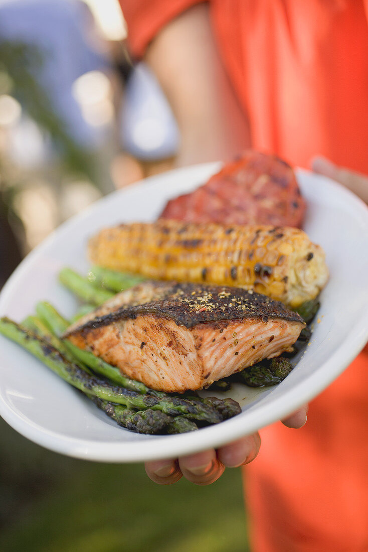 Woman holding plate of grilled salmon, corn on the cob & vegetables