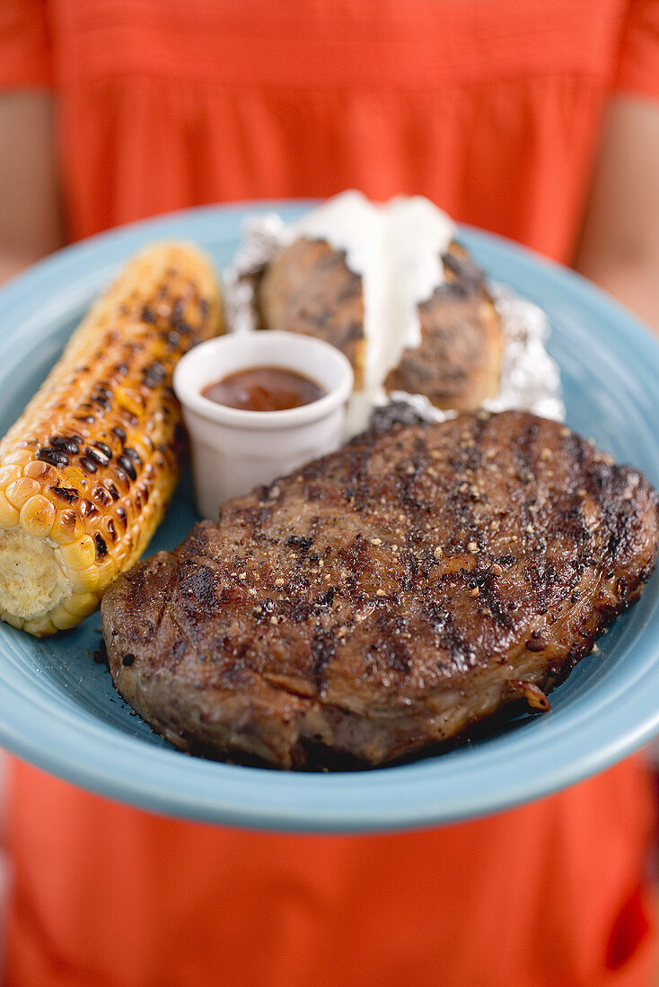 Frau hält Teller mit Grillsteak, Maiskolben, Baked Potatoe