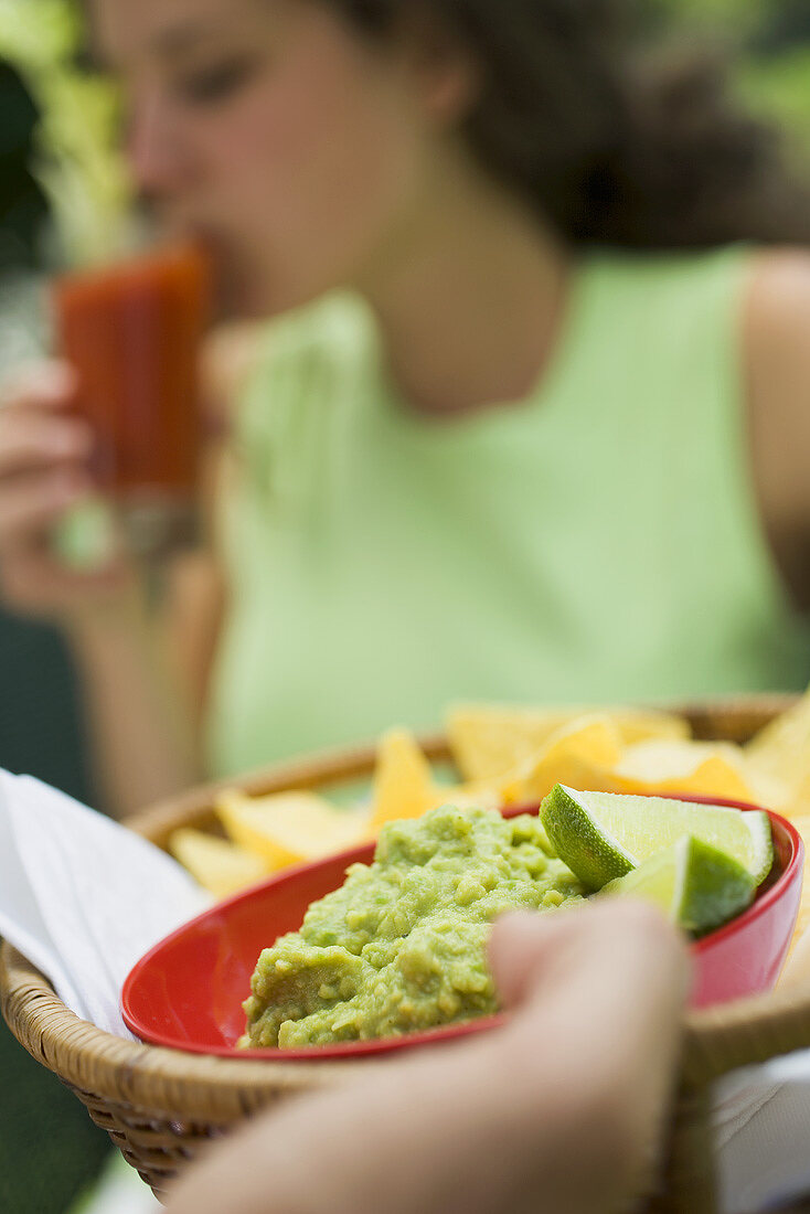 Hand hält Korb mit Guacamole und Chips, Frau im Hintergrund