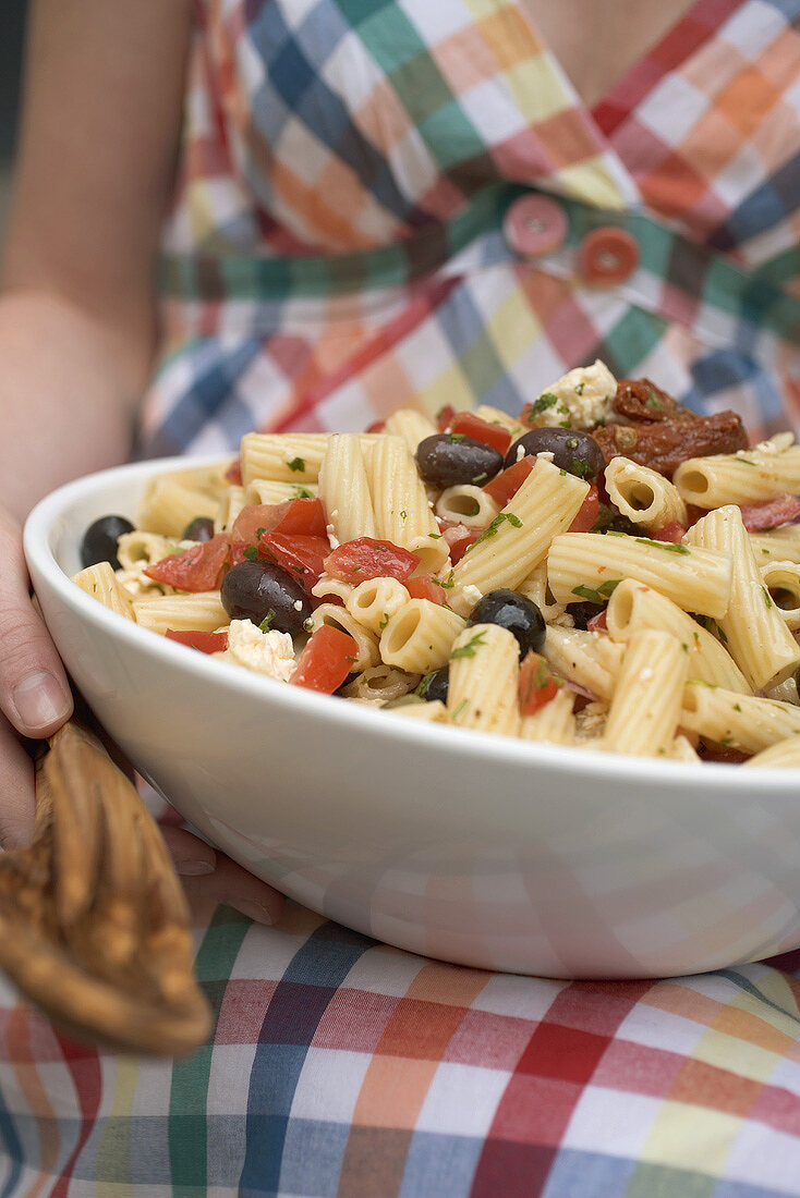 Woman holding large dish of pasta salad with olives & tomatoes