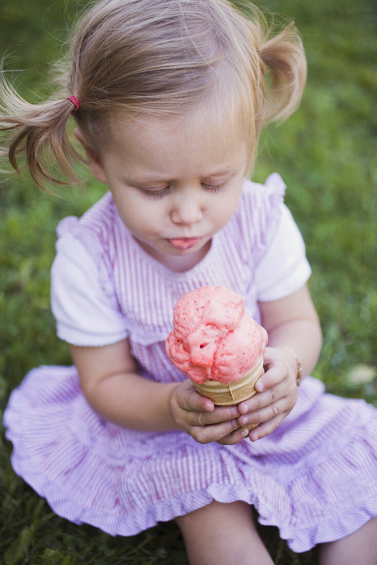 Kleines Mädchen, auf Wiese sitzend, hält Tüte Erdbeereis