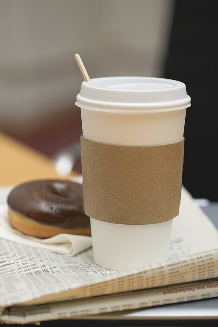 Coffee in paper cup and doughnut on newspaper in office