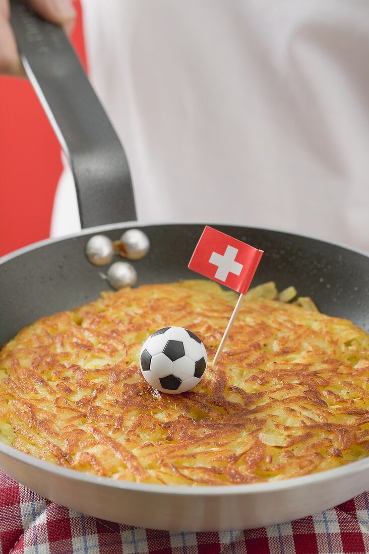 Hand hält Pfanne mit Rösti, Minifussball und Schweizer Flagge