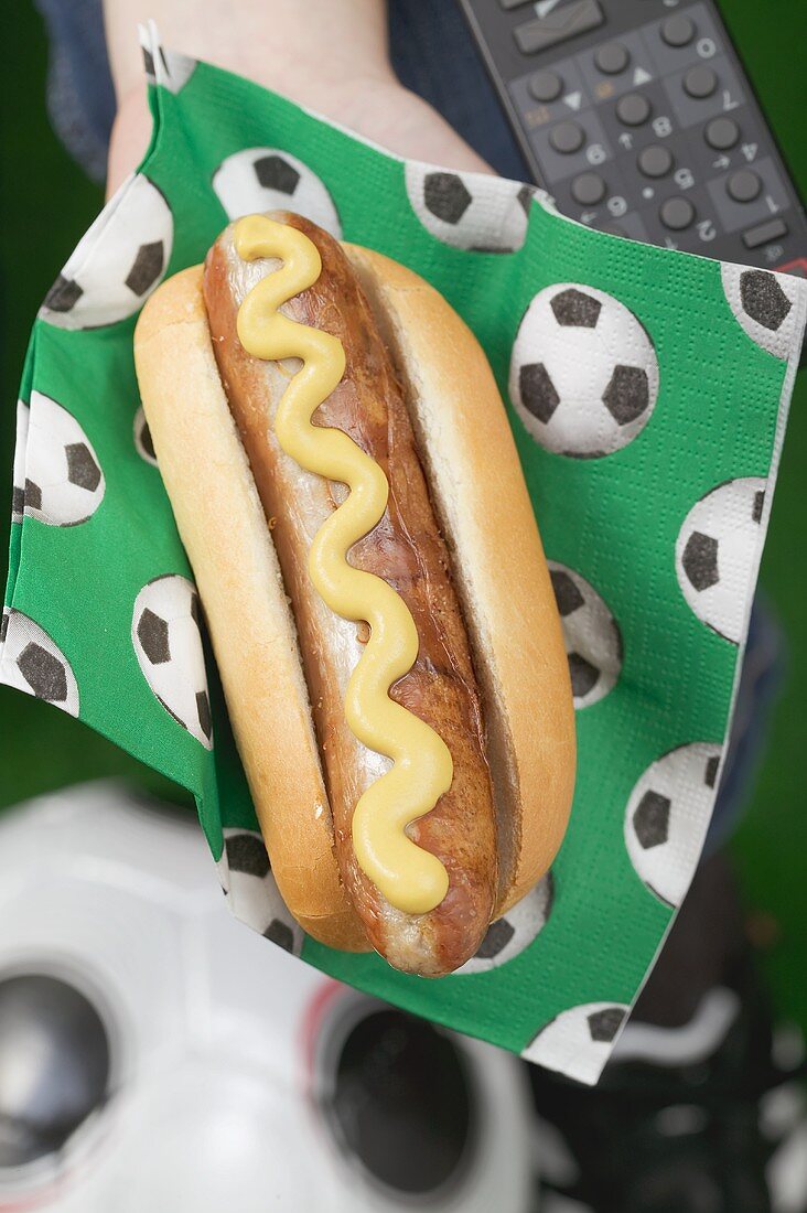Hand hält Hot Dog mit Senf auf Serviette mit Fussballmotiv
