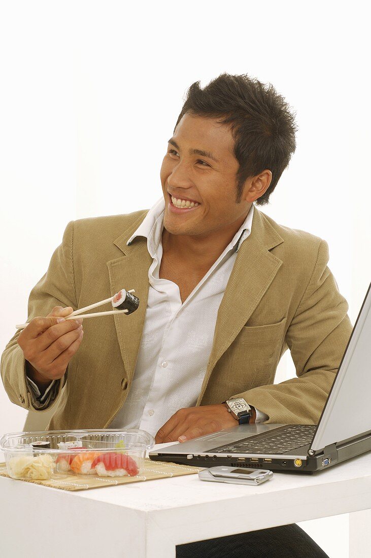 Asian man eating sushi while sitting at laptop