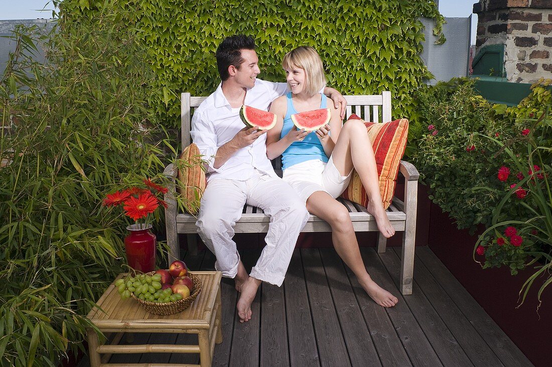 Couple sitting on a bench out of doors & eating watermelon
