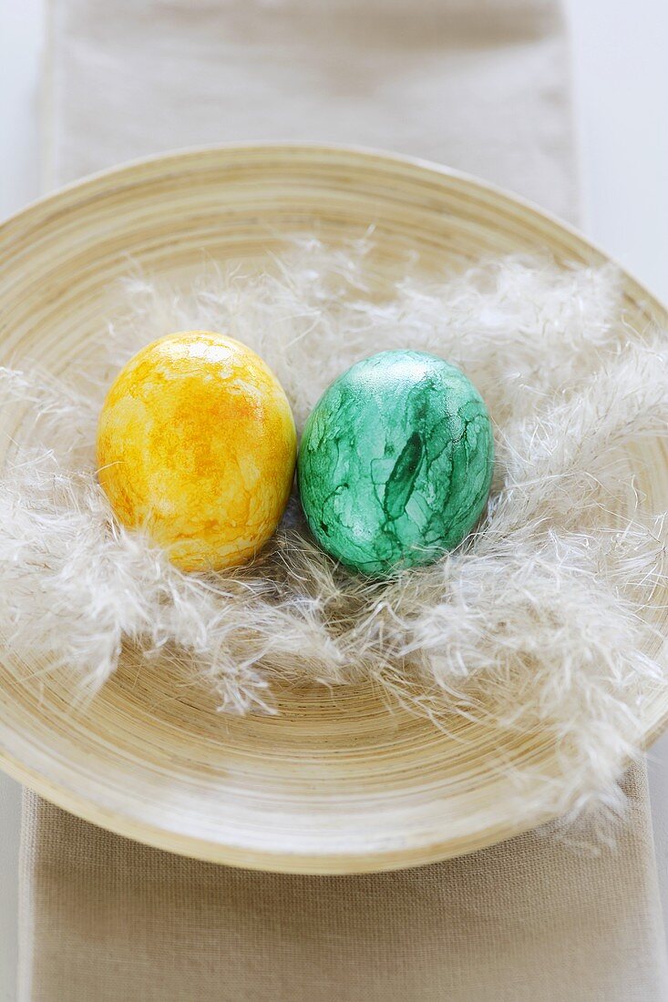Two coloured Easter eggs with feather in wooden bowl