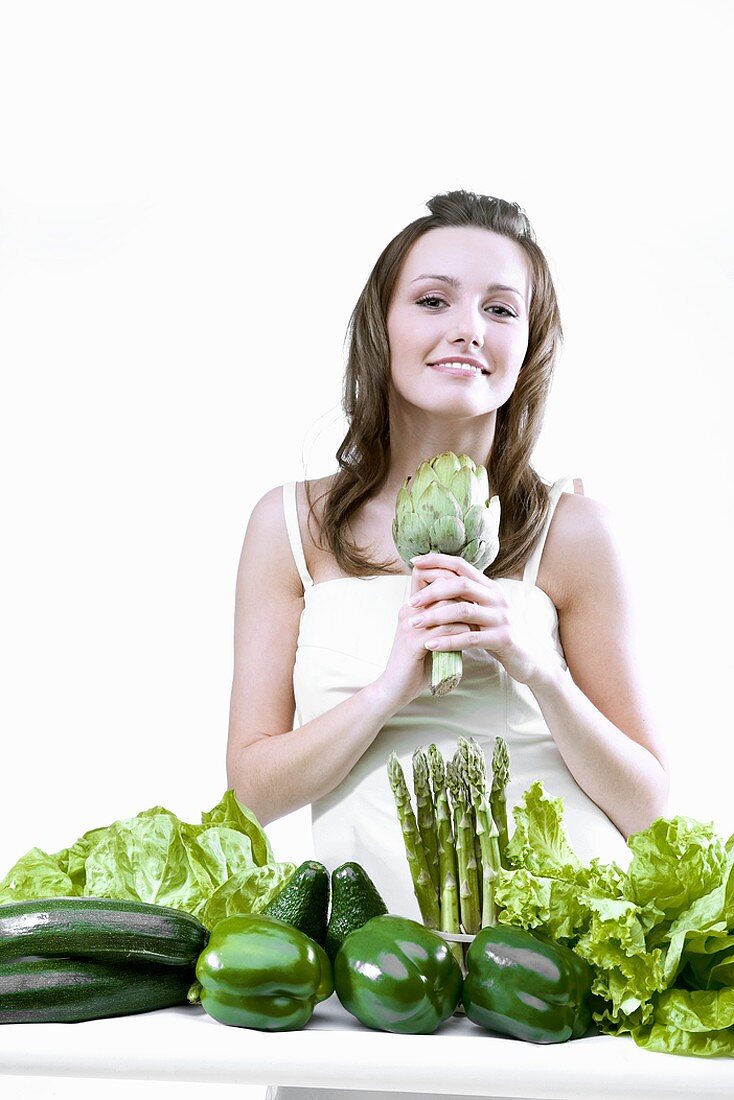 Young woman holding an artichoke, green vegetables in front