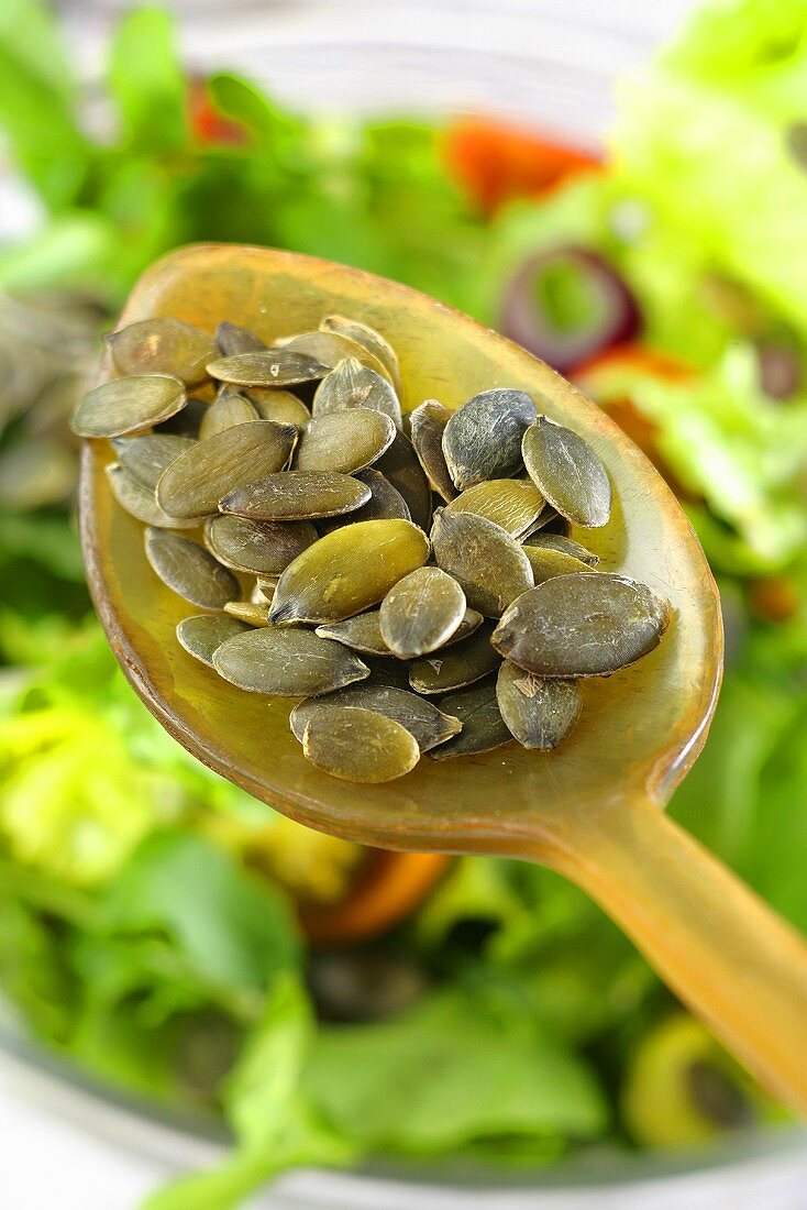Pumpkin seeds on a spoon above a bowl of salad