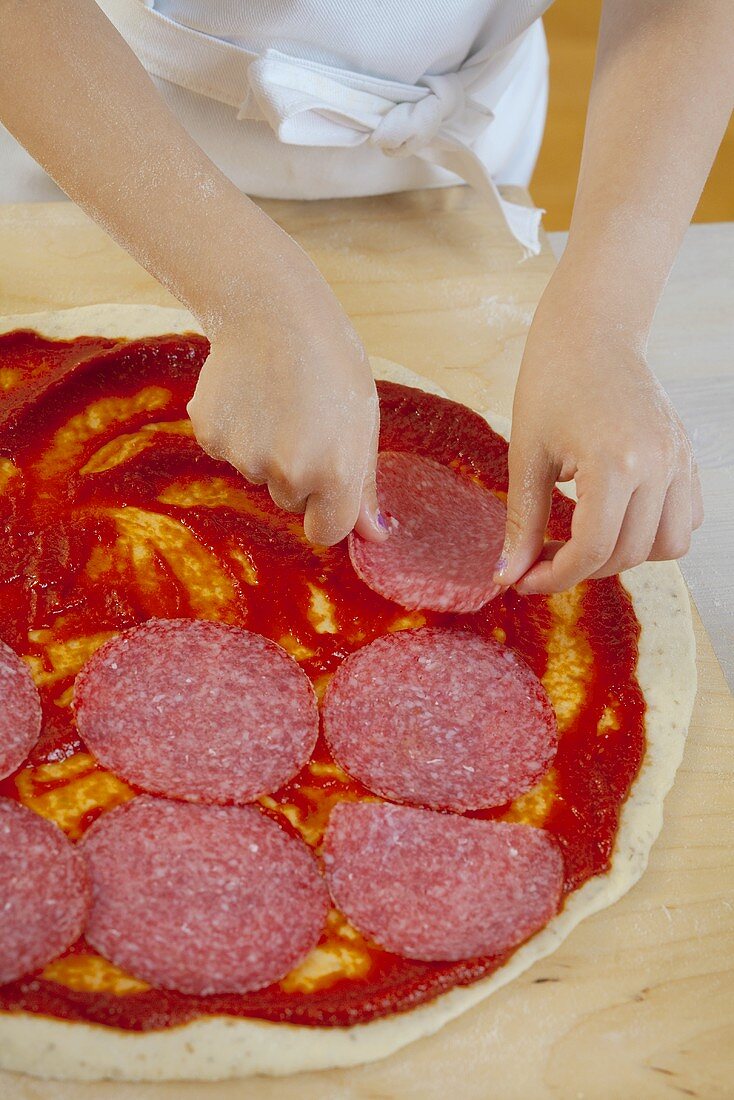 Girl putting slices of salami on pizza