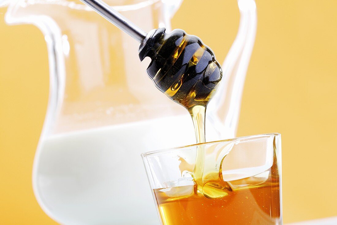 Honig tropft vom Honiglöffel in Glas, Milchkrug