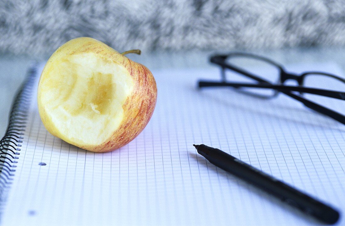 Angebissener Apfel, Brille und Schreibstift auf Notizblock