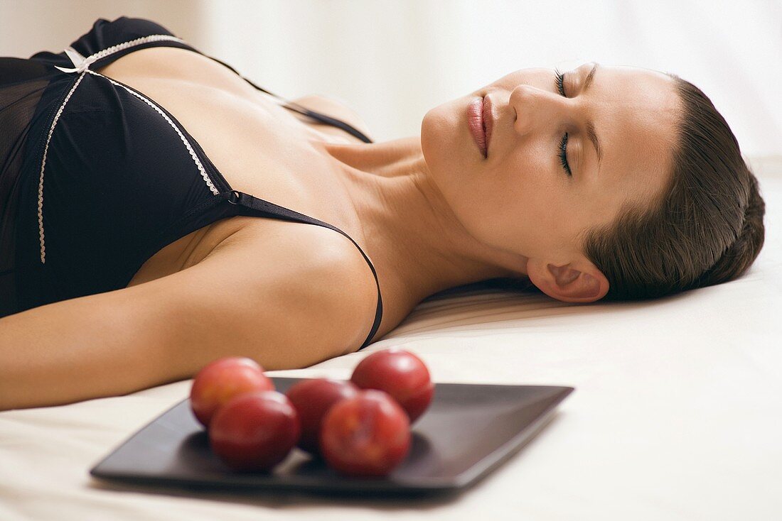 Young woman wearing neglige, relaxing on bed alongside tray with plums