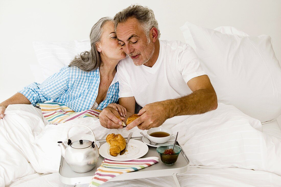 Mature couple sitting on bed with breakfast, women kissing man