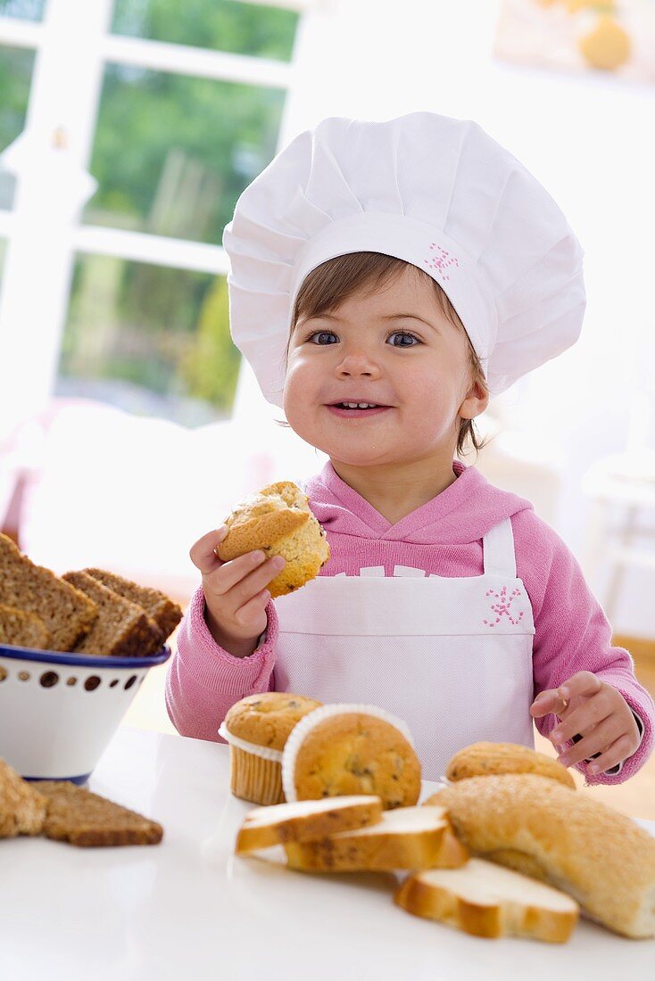 Kleines Mädchen mit Kochmütze sitzt am Tisch vor Muffins